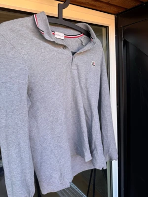 Grå långärmad pikétröja Moncler🙌🤩 - Snygg grå långärmad pikétröja från Moncler med klassisk krage och knappslå. Tröjan har Moncler-logga broderad på bröstet och kragen har röd, vit och marinblå rand. Perfekt för en stilren och avslappnad look.