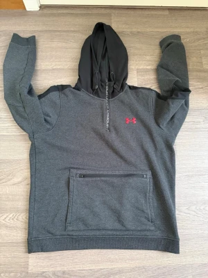 Mörkgrå hoodie från Under Armour - Snygg mörkgrå hoodie från Under Armour med svart huva och dragkedjedetalj på magfickan. Röd logga på bröstet och justerbar huva med snören. Perfekt för en sportig och avslappnad stil.
