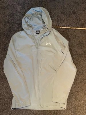 Under Armour - Snygg grå vindjacka från Under Armour i storlek M med luva och dragkedja framtill. Jackan har elastiska muddar vid ärmsluten och en vit logga på bröstet. Tillverkad i lätt och vindtätt material, perfekt för blåsiga dagar.
