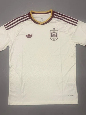 Spanien vm tröja - Vit Spanien fotbollströja från Adidas med bruna detaljer och gula inslag vid kragen. Tröjan har bruna Adidas-ränder på axlarna, broderad landslagssköld och Adidas-logga på bröstet. Tillverkad i lätt och ventilerande material, perfekt för träning eller match.