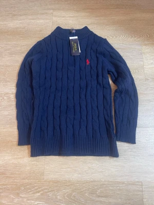 Mörkblå kabelstickad tröja Polo Ralph Lauren - Mörkblå kabelstickad tröja från Polo Ralph Lauren med klassisk röd broderad logga på bröstet. Tröjan har ribbstickade muddar och hög hals. Perfekt för dig som gillar stilren och tidlös design.