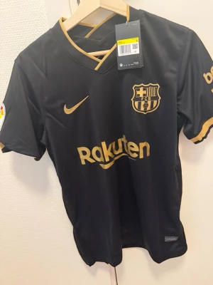 Svart FC Barcelona matchtröja Nike - Snygg svart FC Barcelona matchtröja från Nike med guldiga detaljer och klubbmärke på bröstet. Tröjan har korta ärmar, V-ringning och tryckta loggor. Perfekt för fotbollsträning eller att bära som supporter.