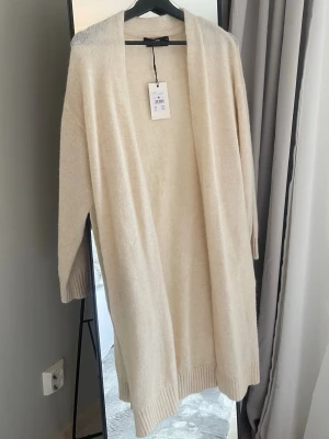 Lång beige kofta från Bik Bok - Helt ny! Lång, öppen beige kofta från Bik Bok i mjuk alpackablandning. Koftan har ribbade muddar och en avslappnad passform, perfekt att slänga över en outfit för extra lager. Enkel och stilren design utan knappar. Skulle ha använt till jul men valde annan outfit, därför är inte ens prislappen borttagen. Köpte för 499kr och säljer för 349kr.
