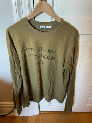  Acne Studios Tröja - Olivgrön långärmad tröja från Acne Studios med tryck 'Acne Studios STOCKHOLM 1996' på bröstet. Tillverkad i mjuk bomull och har en avslappnad passform med rund hals. Perfekt för en chill look