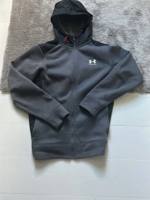 Mörkgrå hoodie från Under Armour - Snygg mörkgrå hoodie från Under Armour med dragkedja och huva. Loggan är tryckt i vitt på bröstet och huvan har röda detaljer på snörena. Tillverkad i mjukt och bekvämt material, perfekt för chill eller träning.