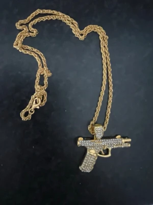 Guldfärgat halsband med pistolhänge - Unikt halsband med en guldfärgad kedja och ett stort pistolformat hänge täckt av glittrande stenar. Kedjan är tvinnad och ger ett coolt intryck, perfekt för dig som vill sticka ut med din stil. Märke ej angivet.