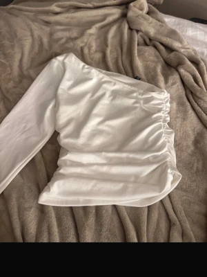 Vit oneshoulder topp med rynk - Trendig vit oneshoulder topp med lång ärm och snygga rynkade detaljer på sidan. Topp i stretchigt material som sitter bekvämt och ger en cool siluett. Perfekt för dig som vill sticka ut med en unik och stilren look. säljs ej längre 