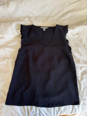 Svart ärmlös topp från H&M - Stilren svart topp från H&M med v-ringning och volangdetaljer vid axlarna. Toppen är ärmlös och har en enkel, rak passform som passar till många olika looks. Perfekt att matcha med jeans eller kjol för en trendig outfit.
