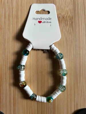 Handgjort armband med pärlor - Snyggt handgjort armband med vita platta pärlor och större glaspärlor i grönt, blått och guld. Armbandet har en modern och fräsch look, perfekt för att addera färg till din stil. Smidig att ta på och av tack vare elastisk tråd.