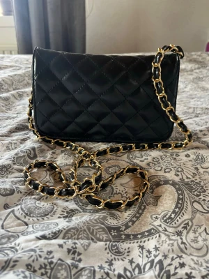 Svart quiltad handväska med kedja - Snygg svart handväska i quiltat mönster med lång axelrem i guld- och svartfärgad kedja. Klassisk rektangulär form och elegant design som ger en lyxig känsla. Perfekt accessoar för att lyfta din outfit.