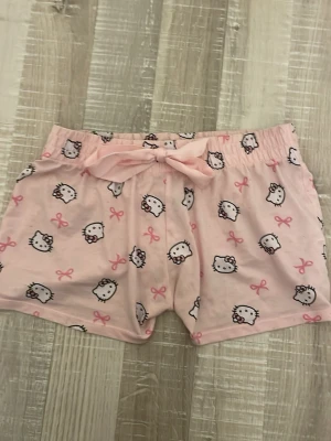 Hello Kitty pyjamasshorts - Söta rosa pyjamasshorts med Hello Kitty-motiv och små rosa rosetter. Shortsen har en mjuk resår i midjan och en dekorativ rosett framtill. Perfekta för mysiga hemmakvällar eller sommarens varma nätter. Tillverkade i mjukt och bekvämt material.