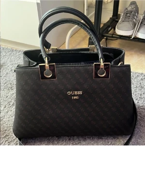 Svart handväska från Guess - Snygg svart handväska från Guess med guldfärgade detaljer och dubbla handtag. Väskan har logomönster, dragkedja upptill och flera fack inuti för smart organisering. Perfekt för dig som vill ha en stilren och trendig accessoar.