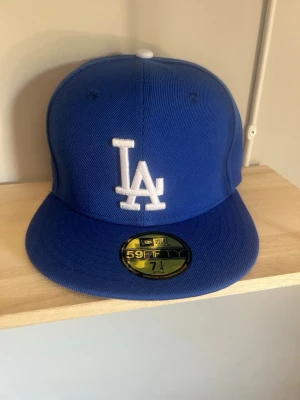 Blå LA Dodgers keps från New Era - Snygg blå 59FIFTY keps från New Era med LA Dodgers logga broderad i vitt framtill. Klassisk flat brim och broderade ventilationshål. Kepsen har New Era-logga på sidan och är tillverkad i ett slitstarkt tyg.