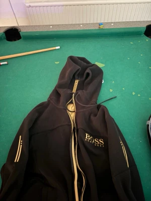 Svart hoodie från Hugo Boss XL - Svart hoodie från Hugo Boss med dragkedja och gulddetaljer. Märkets logga syns på bröstet och längs dragkedjan. Hoodien har huva och ribbade muddar. Perfekt för en stilren och sportig look.