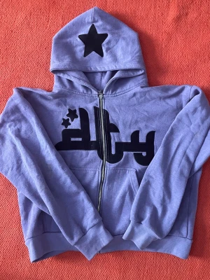 Lila hoodie Divide The Youth - Lila hoodie med dragkedja från Divide The Youth. Stor svart stjärna på huvan och tryck både fram och bak. Mjuk bomullskänsla och cropped passform. Perfekt för dig som gillar streetwear och vill sticka ut med coola detaljer.