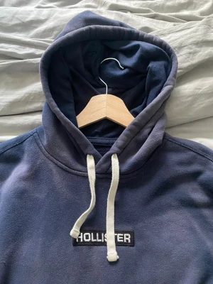 Mörkblå hoodie från Hollister - Snygg mörkblå hoodie från Hollister med vit logga på bröstet och tryck på ärmen. Hoodien har en stor känguruficka framtill och vita dragsnören i huvan. Perfekt för en avslappnad och cool stil.