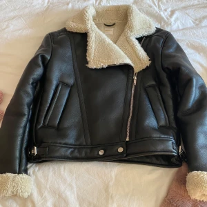 Svart jacka från Gina Tricot - Snygg svart jacka från Gina Tricot med vit fluffig krage och ärmslut. Jackan har en cool asymmetrisk dragkedja, två fickor och bältdetaljer vid midjan. Aldrig använd❣️, perfekt för dig som vill ha en varm look under höst och vinter🩵.