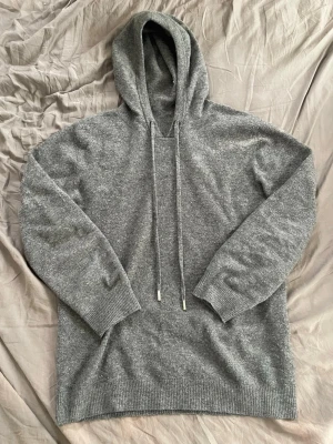 Grå kashmir/merinoull hoodie - Snygg grå hoodie med huva och långa dragsnören. Tillverkad i en mjuk och skön kashmir/merinoull med ribbade muddar vid ärmslut och nederkant. Perfekt för en avslappnad och stilren look.