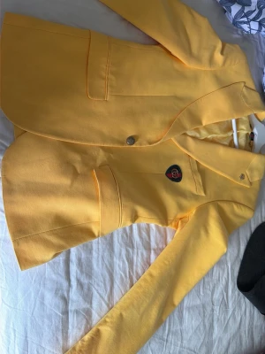 Gul kavaj från Gucci med emblem - Snygg gul kavaj från Gucci med klassiskt GG-emblem på bröstfickan. Kavajen har en stilren design med mjukt tyg och innerfoder. Märket syns tydligt på insidan och detaljerna har en lyxig känsla. Perfekt för dig som vill sticka ut med något unikt och exklusivt.