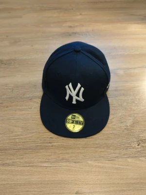 Mörkblå New Era Yankees keps - Snygg mörkblå New Era 59FIFTY keps med klassisk New York Yankees-logga broderad framtill. Kepsen har platt skärm i grått och sticker ut med sin ikoniska stil. Perfekt för dig som vill ha en sportig och trendig look.