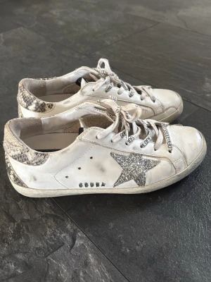 Golden Goose sneakers med glitterstjärna - Säljer ett par vita Golden Goose sneakers med silverglittrig stjärna på sidan och detaljer i ormskinnsmönster på häl och kant. Skorna har text på skosnörena och klassisk låg sneakersmodell. Perfekta för dig som gillar statement-detaljer.