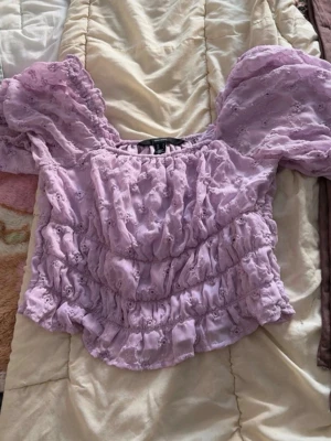 Ljuslila blus med volang och spets - Superfin ljuslila blus med puffiga offshoulder-ärmar och volangdetaljer. Blusen har broderade spetsmönster och är gjord i bomull. Perfekt för dig som gillar romantisk och söt stil.