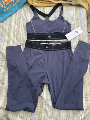 Mörkgrått set från Alo med sport-bh - Snyggt mörkgrått set från Alo med sport-bh och matchande tights. Setet har svarta resårband med vit logga och vita sömdetaljer. Materialet är stretchigt och passar perfekt för en aktiv livsstil.