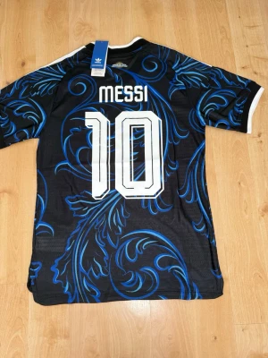 Messi #10 – Player Edition Argentina (storlek M) - Messi #10-tryck. Player Edition Argentina. Storlek M. Helt ny och aldrig använd. Säljs i befintligt skick. Skickas snabbt! 🚚💨  Mått för denna tröja (storlek M): (Uppmätta av leverantör – viss variation kan förekomma) • Halva bröstmåttet: 51 cm • Tröjans längd: 72,5 cm • Ärmbredd: 22,5 cm • Axelbredd: 43 cm • Ärmlängd: 39 cm • Manschettbredd: 18 cm  🔥 Player Edition-modell – tajtare passform än vanliga supportertröjor. 🇦🇷 Argentina landslag.