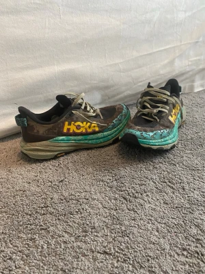 Hoka speed goat 6 löparskor  - Säljer ett par Hoka löparskor i svart och brunt med turkos och gul detalj på sulan. Skorna har snörning och är tillverkade i mesh och syntetmaterial för bra andningsförmåga och komfort. Perfekta för dig som gillar att springa långt och vill ha bra dämpning.
