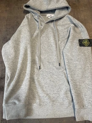 Grå hoodie från Stone Island - Snygg grå hoodie från Stone Island med klassisk huva och dragsnören. Ikonisk logotyp-patch på vänster ärm. Tillverkad i mjuk bomullsblandning och har ribbade muddar vid ärmslut och nederkant. Perfekt för en avslappnad streetwear-look.