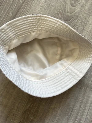 Beige bucket hat med rutigt mönster - Säljer en beige bucket hat med klassiskt rutigt mönster i svart, vitt och rött. Hatten har bred brätte och är tillverkad i ett lätt tyg, perfekt för en trendig streetwear-look. Insidan är helvit och mjuk för extra komfort.