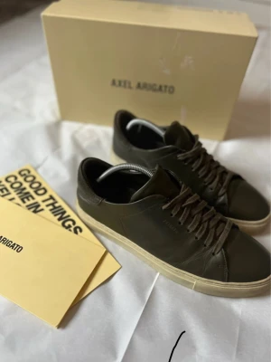 Axel Arigato gröna lädersneakers - Stilrena sneakers från Axel Arigato i mörkgrönt läder med matchande gröna skosnören och vit sula. Klassisk låg modell med minimalistisk design och diskret logga på sidan. Perfekta för dig som gillar en clean och modern look.
