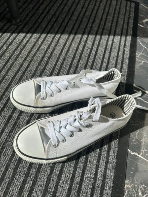 Vita låga tygsneakers med ränder - Snygga vita sneakers i tyg med klassisk låg modell och vita snören. Skorna har svart-vit randigt foder och en vit sula med svart rand. Perfekta för en clean och stilren look till vardagsoutfiten.