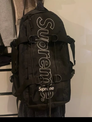 Supreme svart ryggsäck FW18 - Säljer en svart ryggsäck från Supreme FW18 med stor vit logga längs framsidan. Ryggsäcken har flera justerbara remmar, dragkedjefack och robust design. Perfekt för dig som vill ha streetwear-vibe och mycket plats för grejer.