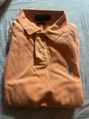 Orange pikétröja från Polo Ralph Lauren - Säljer en klassisk orange pikétröja från Polo Ralph Lauren med blå logga på bröstet. Tröjan har korta ärmar, krage och knappar framtill. Perfekt för en avslappnad och stilren look.