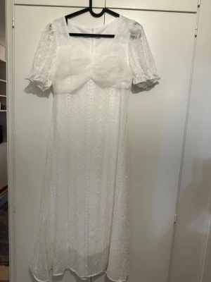 Vit lång spetsklänning med puffärm - Romantisk lång vit klänning med spetsdetaljer och puffärmar. Klänningen har en markerad byst med voluminös tyllrosett framtill och transparenta ärmar. Perfekt för dig som gillar vintageinspirerad stil och vill sticka ut med en unik look.