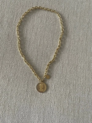 Guldfärgat halsband med myntberlock - Snyggt halsband i guldfärgad metall med grova länkar och en rund myntberlock med profilmotiv. Kedjan har även en liten rund detalj nära berlocken. Perfekt accessoar för att lyfta din outfit.