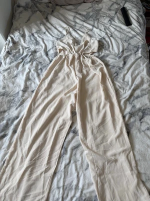Beige jumpsuit med smala axelband - Snygg beige jumpsuit med smala axelband och v-ringning. Modellen har markerad midja med resår och vida ben. Materialet är tunt och mjukt, perfekt för varma dagar. Cut-out detaljer vid midjan ger en trendig look.
