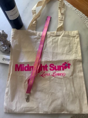 Merch  Zara Larsson Midnight sun tour - Beige shoppingväska i tyg med trycket 'Midnight Sun Zara Larsson' i rosa. Väskan har långa handtag och är perfekt för att bära med sig allt man behöver. Snygg och enkel design med färgglad text framtill. Denna väska samt nyckelring fick man som VIP så går inte köpa som andra merch, vilket gör dem mer unika! Nyckelringen är rosa med gul text. 