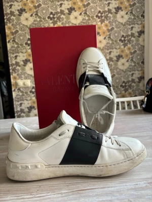Valentino vita sneakers med svart detalj - Snygga vita sneakers från Valentino med bred svart läderdetalj över mitten och klassisk logga på plösen. Skorna har rund tå, vita snören och diskreta nitar längs sulan. Perfekta för dig som gillar stilrena och exklusiva sneakers. Kan så klart tvätta innan ni köper. Har kvitto.