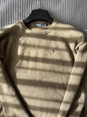 Grön stickad Ralph Lauren tröja  - En stilren beige stickad tröja med rund halsringning och långa ärmar. Tröjan har en enkel och klassisk design som passar perfekt till många olika outfits. Materialet ser mjukt och bekvämt ut, perfekt för kyligare dagar.