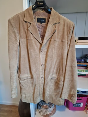 Beige kavaj från Selected i mocka - Stilren beige kavaj från Selected i mjuk mocka. Klassisk passform med tre knappar framtill, två fickor och snyggt skuren krage. Perfekt för dig som vill ha en tidlös och trendig look.