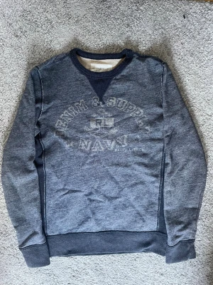Grå sweatshirt från Denim & Supply - Mörkgrå sweatshirt från Denim & Supply Ralph Lauren med tryckt text och ankare på bröstet. Tröjan har ribbade muddar vid ärmslut och nederkant samt en klassisk crewneck. Tillverkad i bomull för en skön känsla.