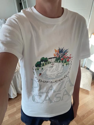 Vit t-shirt med Plaza Casa-tryck - Vit t-shirt med rund hals och tryck framtill som föreställer en villa med palmer och blommor samt texten 'PLAZA CASA' i stora bokstäver. T-shirten har en klassisk passform och är tillverkad i mjukt bomullsmaterial. Perfekt för en avslappnad och trendig look.