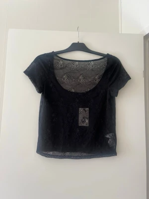 Svart spetsblus med kort ärm - Snygg svart blus i transparent spets med blommigt mönster och korta ärmar. Perfekt för lager-på-lager och ger en cool vibe till din outfit. Djup rund halsringning och lätt, luftig känsla.