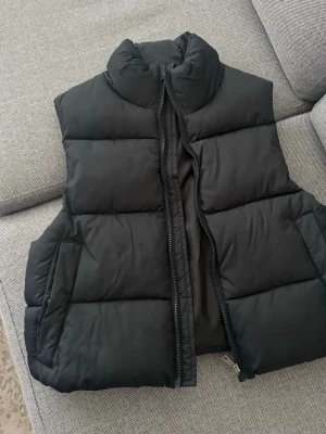 Svart puffad dunväst med hög krage - Snygg svart dunväst med puffad design och hög krage. Västen har dragkedja framtill och två sidofickor. Perfekt för lager-på-lager och streetstyle. Materialet är quiltat och ger en modern look.