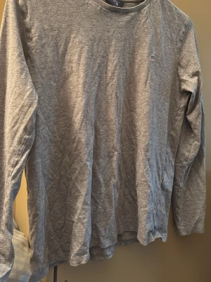Grå långärmad tröja Gant - En stilren grå långärmad tröja från Gant med rund halsringning och liten broderad logga på bröstet. Tillverkad i mjuk bomull, perfekt för en avslappnad och enkel look.
