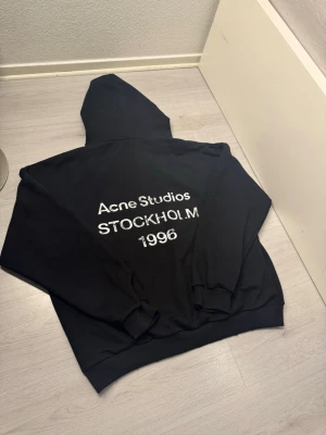 Svart hoodie Acne Studios Stockholm - Svart hoodie från Acne Studios med vit text på ryggen där det står 'Acne Studios STOCKHOLM 1996'. Oversized passform med huva och ribbade muddar. Perfekt för dig som gillar stilrena och trendiga plagg med streetkänsla. Helt oanvänd. Storlek L. Fraktar samma dag eller dagen efter. Från axel till axel 51 cm. Axle till midjan 68 cm :)