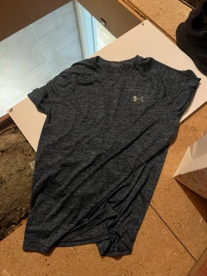 Mörkblå Under Armour t-shirt - Snygg mörkblå t-shirt från Under Armour med diskret logga på bröstet. T-shirten är kortärmad och har en sportig, melerad look. Perfekt för dig som gillar en stilren och bekväm stil. Materialet känns lätt och andas bra.
