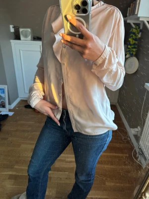 Ljusrosa blus med volangdetalj - Säljer en ljusrosa blus med volang längs knappraden och långa ärmar med resår vid ärmslut. Skjortan har en mjuk, glansig yta och klassisk krage. Perfekt för dig som gillar en feminin och trendig look.